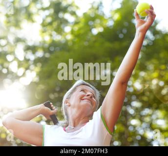 Bereit für den perfekten Aufschlag - Tennis. Lächelnde Seniorin, die ihre Arme hebt, um sich auf einen Tennisaufschlag vorzubereiten. Stockfoto