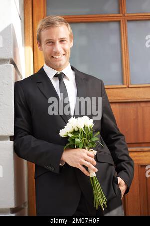 Selbstbewusst in der Liebe. Porträt eines gutaussehenden Mannes, der sich an eine Wand lehnt, einen Haufen weißer Rosen hält und lächelt. Stockfoto