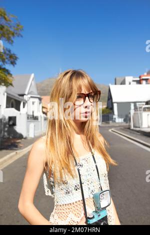 Mädchen in der Stadt. Ein attraktives junges Mädchen mit einer Oldtimer-Kamera, das mitten auf einer Straße in einer Wohngegend steht. Stockfoto