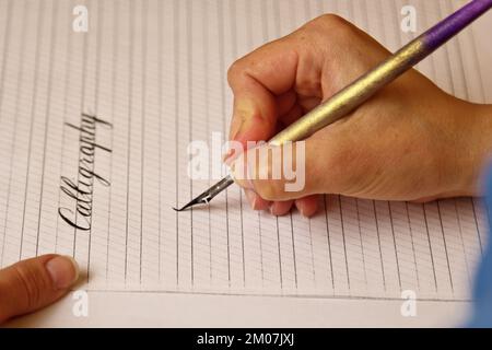 Weibliche Hand schreibt mit dem Tintenstift das Wort Kalligrafie auf ein Papierblatt mit Streifen. Schreibwaren auf dem Schreibtisch aus nächster Nähe. Rechtschreibunterricht und ca. Stockfoto