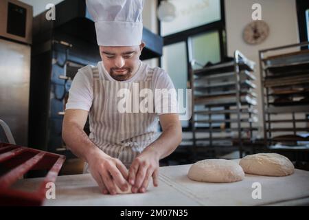 Junger Bäcker mit Down-Syndrom, der Gebäck in der Bäckerei herstellt. Konzept der gesellschaftlichen Integration von Menschen mit Behinderungen. Stockfoto