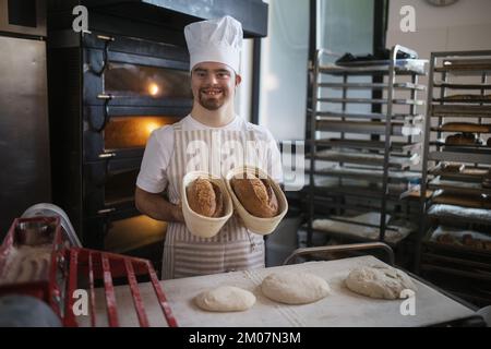 Junger Bäcker mit Down-Syndrom, der Gebäck in der Bäckerei herstellt. Konzept der gesellschaftlichen Integration von Menschen mit Behinderungen. Stockfoto