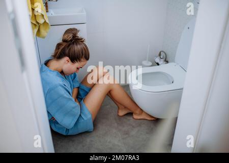 Junge kranke Frau, die auf dem Boden neben der Toilette sitzt. Stockfoto