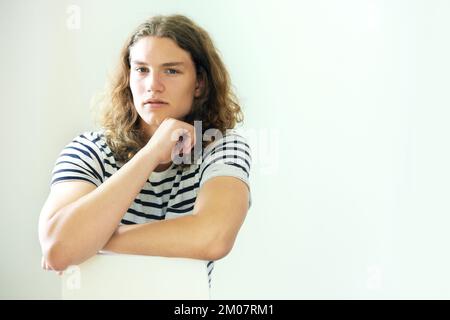 Ich denke darüber nach. Lässiger junger Mann mit langen, lockigen Haaren, der sich auf einen Rückwärtsstuhl lehnt. Stockfoto