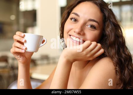 Eine glückliche Frau, die eine Kaffeetasse hält, sieht dich in einer Bar sitzen Stockfoto