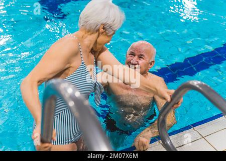 Ältere sportliche weiße Großmutter im gestreiften Badeanzug, die ihren Mann ansieht, während sie langsam im Poolwasser auftaucht. Positive und aktive Freizeit. Hochwertiges Foto Stockfoto