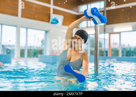 Aqua-Fitnesskonzept. Engagierte glückliche Rentnerin in ihren späten 60s Jahren, die mit Schwimmbadbojen trainierte, während sie auf halbem Weg in Wasser eingetaucht war. Verschwommenes Innenleben des Swimmingpools im Hintergrund. Hochwertiges Foto Stockfoto