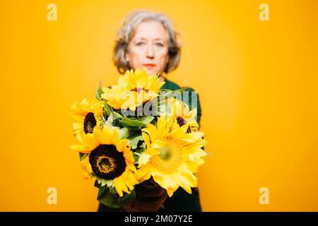 Seniorin mit einem Sonnenblumenstrauß auf gelbem Hintergrund Stockfoto