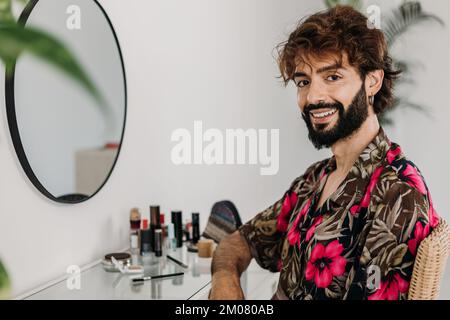 Fröhlicher, mittelgroßer, schwuler Mann, der vor der Kamera lächelt, sitzt am Frisiertisch Stockfoto