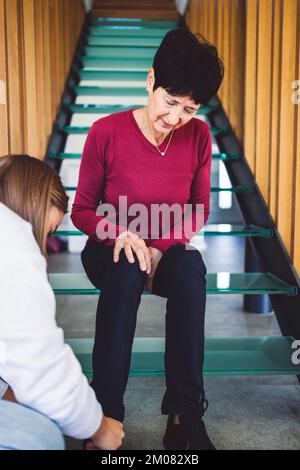 Ein vertikales Foto einer Seniorin, die auf einer gläsernen Treppe sitzt, während eine unbekannte Krankenschwester ihr hilft, Schuhe anzuziehen Stockfoto