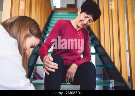 Seniorin sitzt geduldig auf der Treppe und wartet darauf, dass die Krankenschwester ihre Schuhe zubindet Stockfoto