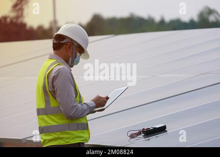 Ein junger asiatischer Ingenieur, der einen Helm und eine weiße Maske trägt, prüft die Sauberkeit von Solarmodulen – Konzept für erneuerbare Energie Stockfoto