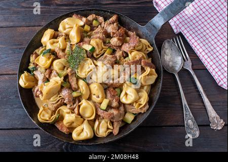 Tortellini mit Fleisch-, Zucchini- und Sahne-Sauce in einer rustikalen Eisenpfanne, isoliert auf einem Holztisch. Draufsicht Stockfoto