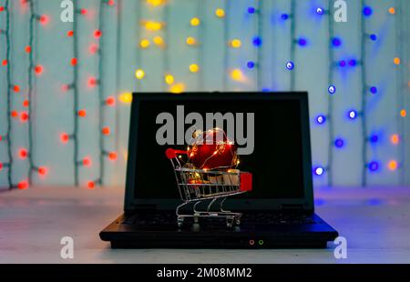 Einkaufswagen mit großem weihnachtsball und Girlanden auf der Tastatur des Laptops. Hintergrund - farbige Girlande. Stockfoto