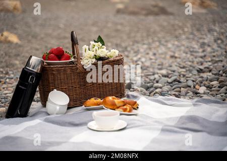 Konzept für Speisen, Urlaub und Feiern - geschlossener Picknickkorb mit Thermoskanne mit Tee, Brötchen, Erdbeeren, Blumen am Strand Stockfoto