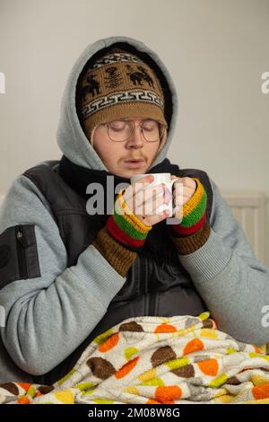 Ein junger Mann, eingewickelt in schweren Mantel, Handschuhe und Hut, der sich mit einer heißen Tasse Tee aufwärmt Stockfoto