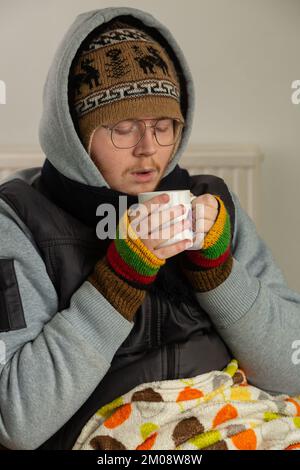 Ein junger Mann, eingewickelt in schweren Mantel, Handschuhe und Hut, der sich mit einer heißen Tasse Tee aufwärmt Stockfoto