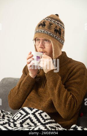 Ein junger Mann mit Wollmütze und Pullover, der einen Kräutertee trinkt Stockfoto