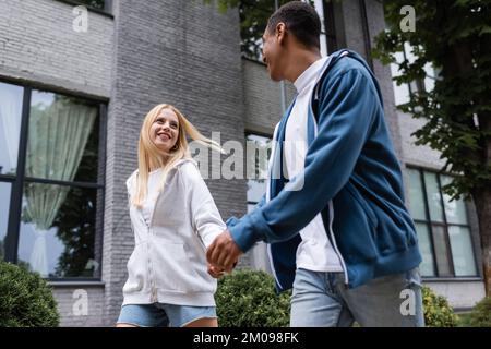 Ein multiethnisches Paar in Kapuzenpullis, das auf der Straße spaziert, Händchen hält und sich anschaut Stockfoto