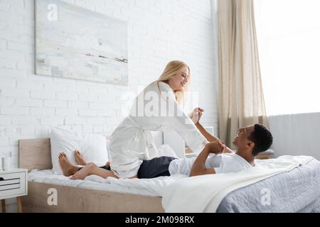 Fröhliche blonde Frau im weißen Bademantel, die Spaß mit einem afroamerikanischen Freund hat, der auf dem Bett liegt Stockfoto