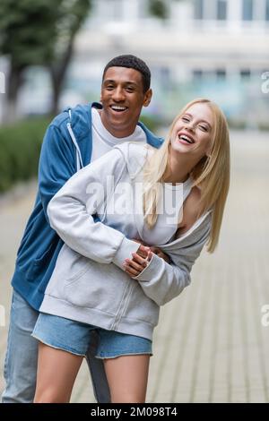 Eine glückliche blonde Frau mit einem afroamerikanischen Freund in Hoodies, die auf der verschwommenen Straße in die Kamera schaut Stockfoto