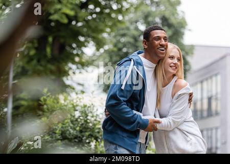 Ein fröhliches gemischtrassiges Paar in Hoodies, das sich umarmt und auf die grüne und verschwommene Straße blickt Stockfoto