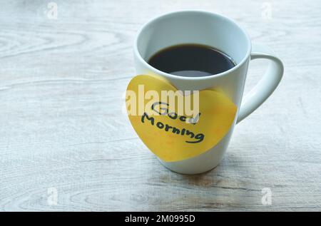 Schwarzer Kaffeetassenstab Good Morning Nachricht in gelbem Herzpapier auf dem Tisch Stockfoto