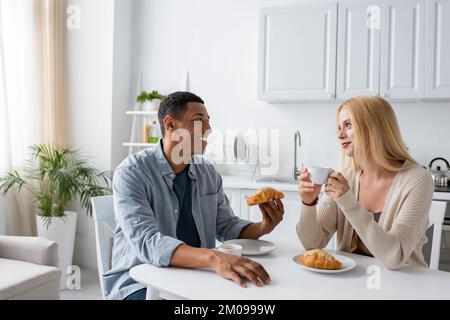 Glückliches gemischtrassiges Paar, das sich beim Frühstück mit Kaffee und leckeren Croissants anschaute Stockfoto