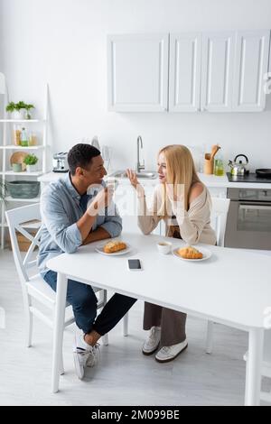 Glückliches gemischtes Paar, das sich in der Nähe von Kaffeetassen und Croissants unterhielt, während es in der Küche frühstückte Stockfoto