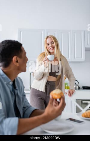 Eine hübsche blonde Frau mit Kaffeetasse, die einen verschwommenen afroamerikanischen Freund beim Frühstück in der Küche ansieht Stockfoto