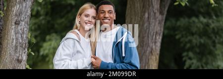 Positives gemischtrassiges Paar in Kapuzen, lächelnd und händchenhaltend im Park, Banner Stockfoto