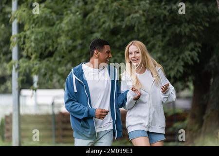 Aufgeregte blonde Frau, die sich im Park mit einem afroamerikanischen Freund unterhalten hat Stockfoto