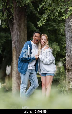 Fröhlicher afroamerikanischer Mann und junge blonde Frau in Kapuzenpullis, die Händchen hält und im Park auf die Kamera schaut Stockfoto