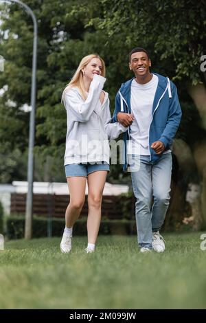 Ein vielsprachiges, lächelndes Paar, das im Park auf dem Rasen spaziert und Händchen hält Stockfoto