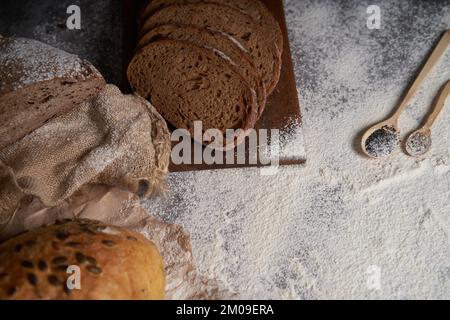 Draufsicht auf verschiedene Brotsorten, geschnitten und ganz, Mehl, zerknittertes Papier und Latte auf einem Holztisch. Großer Winkel. Stockfoto