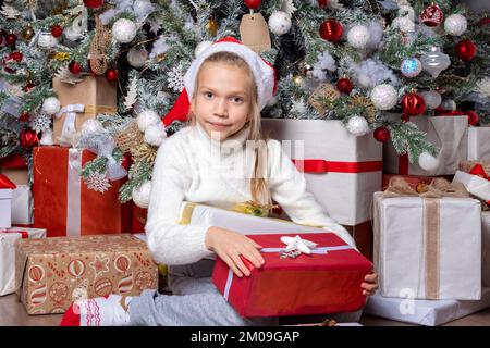 Süßes glückliches Kind mit weihnachtsmannmütze mit einem Überraschungsgeschenk auf dem Hintergrund eines dekorierten Weihnachtsbaums. Zufriedenes Mädchen bekam Weihnachten oder Neujahr Stockfoto