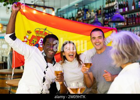 Fröhliche internationale Fußballfans winken mit spanischer Flagge und trinken Bier in der Sportbar Stockfoto