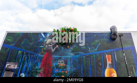 Große Wandgemälde einer Frau in einem leuchtend roten Kleid, die zu lebensechten Plastikblumen neigt, vor einem leuchtend blauen Himmel, auf einer Straße in Shoreditch, E1. Stockfoto