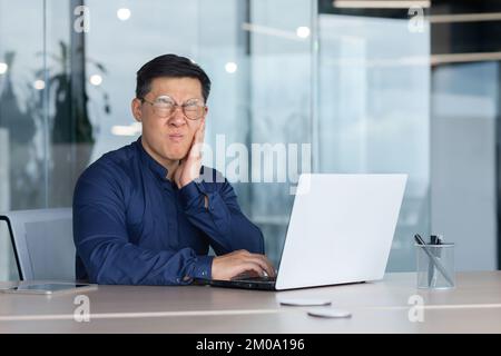 Asiatischer Kranker im Büro, Geschäftsmann hat Zahnschmerzen, Mann sitzt im Büro im Gebäude und arbeitet mit einem Laptop auf der Arbeit, Boss im Hemd neben dem Fenster verärgert. Stockfoto