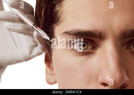 Porträt von jungen Männern, Männchen und kosmetologischen Händen, die über weißem Hintergrund isolierte Augen-Anti-Falten-Injektionen machen. Das Konzept der männlichen Schönheit Stockfoto