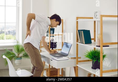 Ein Mann, der an einem Laptop arbeitet, fühlt Rückenschmerzen, steht auf und massiert sich den Rücken, um sich zu entspannen. Stockfoto
