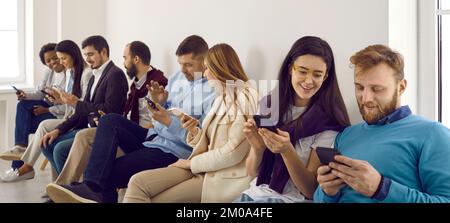 Eine Gruppe glücklicher junger Männer und Frauen, die im Büro sitzen und ihre Smartphones benutzen Stockfoto