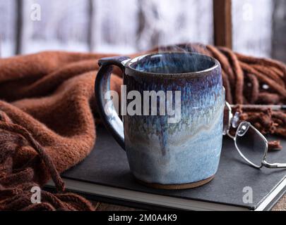 Dampfgaren einer heißen Tasse Kaffee oder Tee an einem Fenster mit verschneitem Winterhintergrund Stockfoto