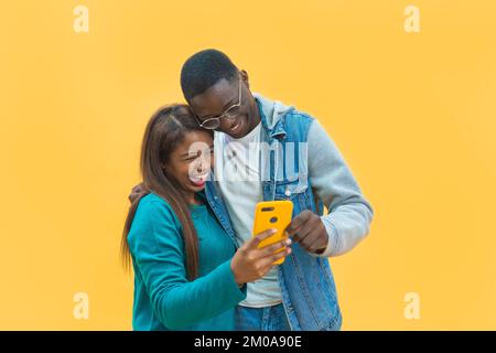 Aufgeregtes schwarzes Paar, das auf einen gelben Studiohintergrund mit Handy-Bildschirm schaut Stockfoto