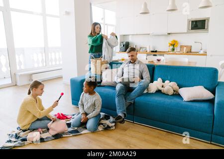 Glückliche Familie zu Hause. Mutter macht Hausarbeit, Vater arbeitet an einem Tablet, Töchter spielen zusammen Stockfoto