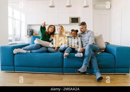 Glückliche Familienmutter, Vater und zwei Töchter sitzen auf dem Sofa und sehen zu Hause fern. Einführungskonzept Stockfoto
