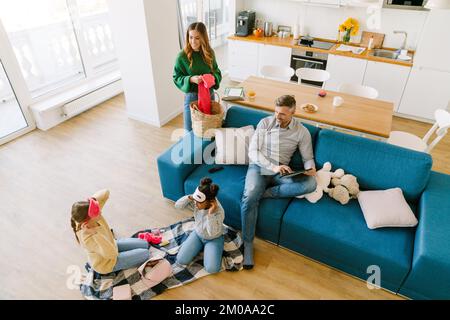 Glückliche Familie zu Hause. Mutter macht Hausarbeit, Vater arbeitet an einem Tablet, Töchter spielen zusammen Stockfoto