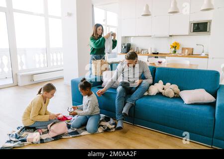 Glückliche Familie zu Hause. Mutter macht Hausarbeit, Vater arbeitet an einem Tablet, Töchter spielen zusammen Stockfoto
