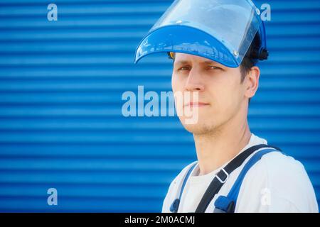 Porträt eines jungen Arbeiters im Schutzhelm auf blauem Industriehintergrund mit Platz zum Kopieren. Kaukasischer Arbeiter selbstbewusster Blick blickt direkt in die Kamera. Stockfoto