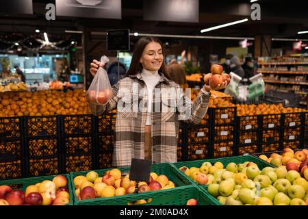 Eine Frau in der Obstabteilung eines Supermarkts kauft Äpfel und überlegt, ob sie eine Plastiktüte verwenden soll Stockfoto